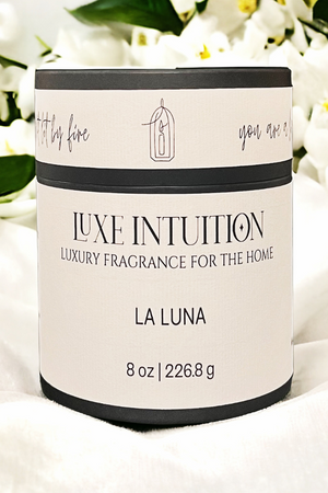 la luna candle in black tube package sitting on white blanket with white flowers in the background.