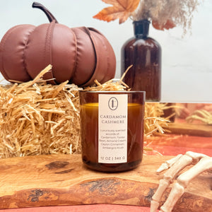 Woodwick Candle labeled 'Cardamom Cashmere' in an amber vessel with decorative pumpkins and hay in the background