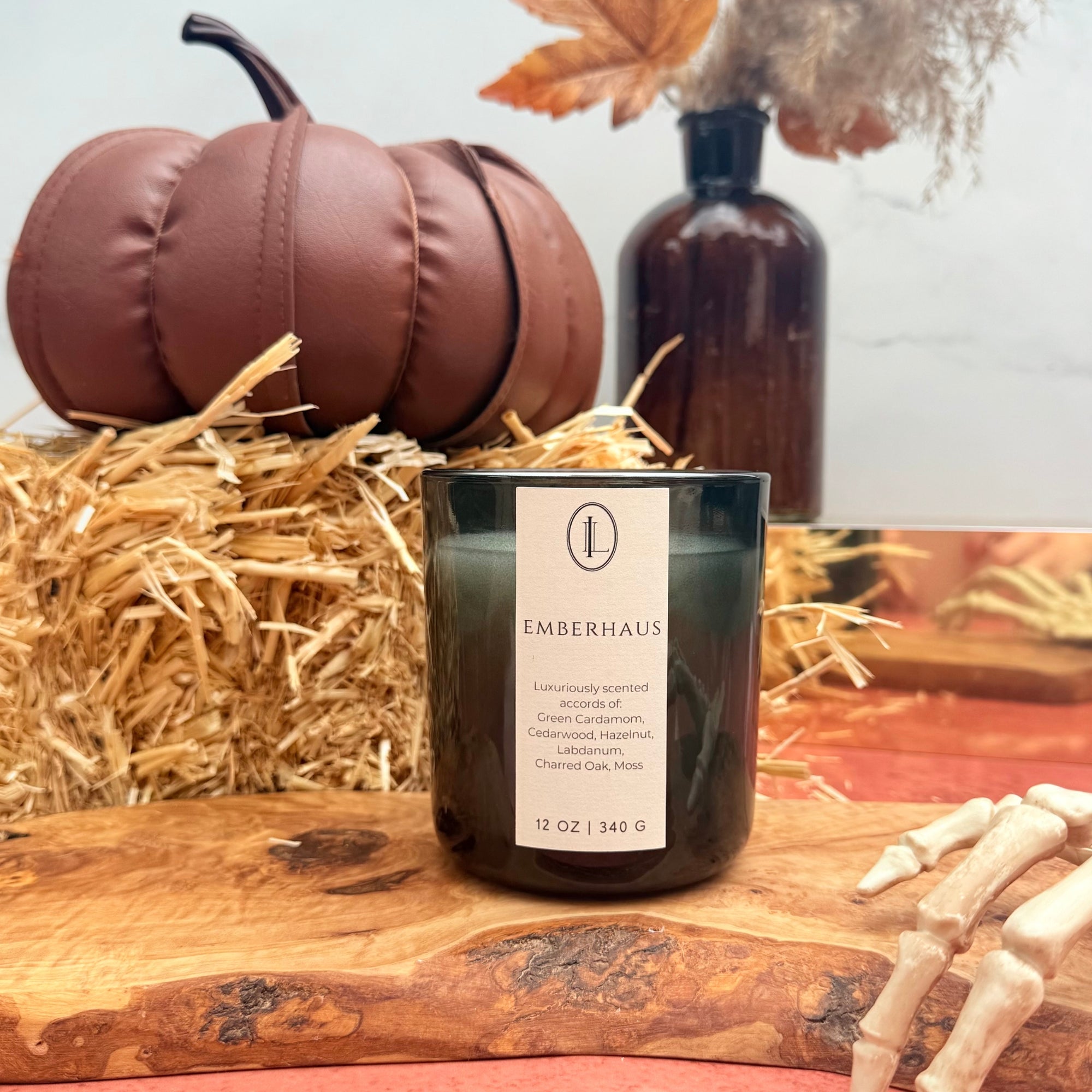 woodwick candle labeled Emberhaus in a translucent black vessel with ay and a pumpkin in the background.